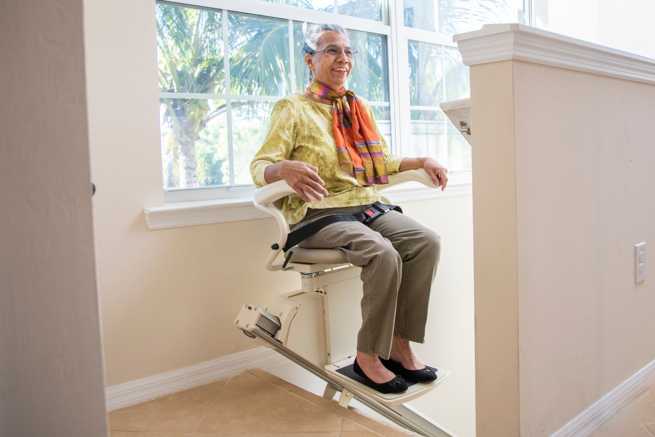 An elderly woman rides a stairlift at home, wearing a yellow top, gray pants, and a colorful scarf. She is smiling and looking out the window beside her.