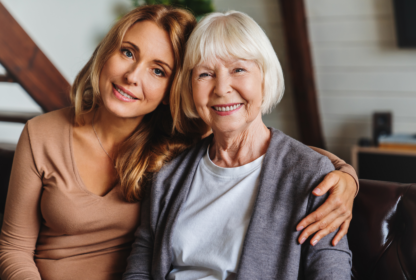 Two women, one younger with long blonde hair and the other older with short white hair, sit closely together on a couch, smiling at the camera in a cozy living room.