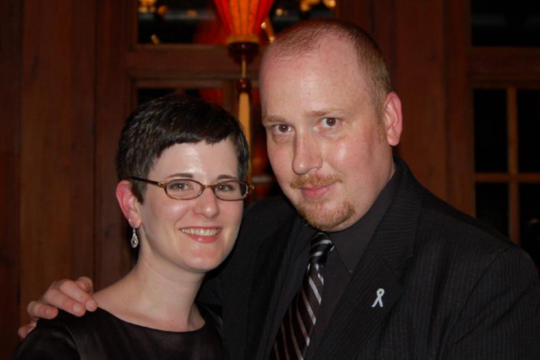 A man and woman pose together, smiling at the camera. The man wears a suit and tie, while the woman has short hair and glasses.