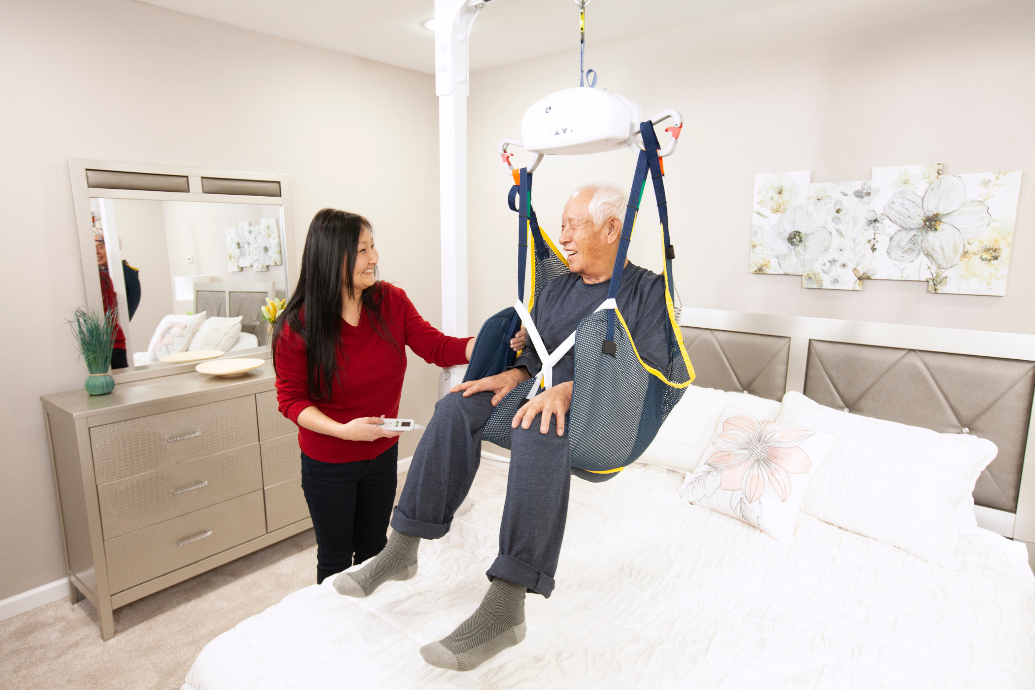 A caregiver assists an elderly man using a ceiling lift above a bed in a bedroom setting. The man is securely seated in the lift, while the caregiver holds a control.