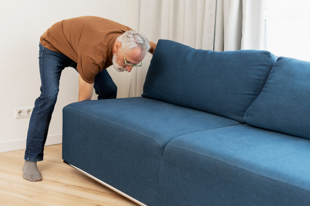 A person in a brown shirt adjusts a blue sofa in a room with light-colored flooring and white curtains.