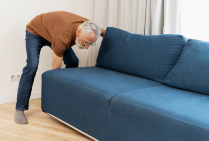 A person in a brown shirt adjusts a blue sofa in a room with light-colored flooring and white curtains.