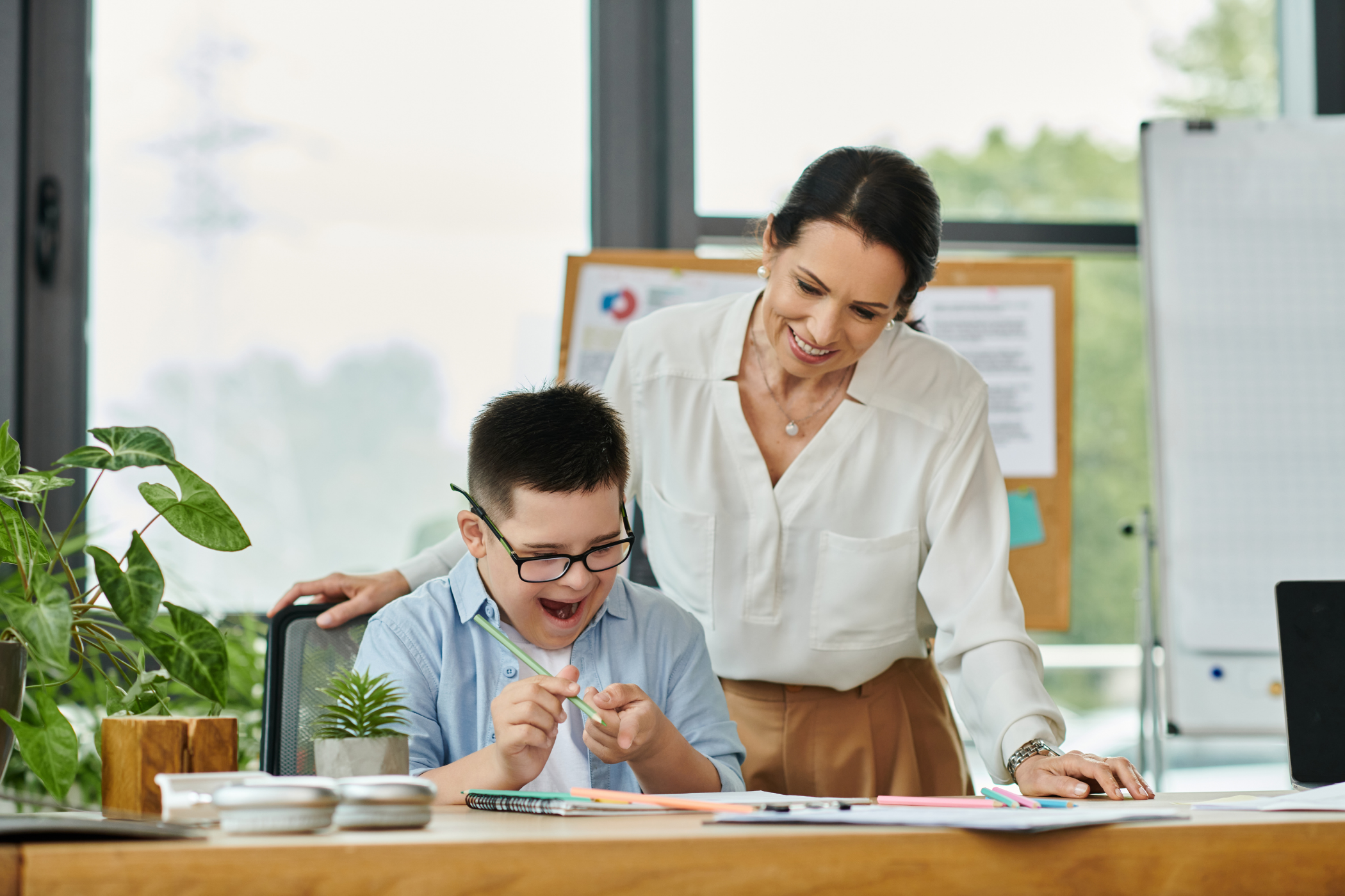 A teacher smiles while assisting an enthusiastic student with his work at a desk, surrounded by plants and stationery.