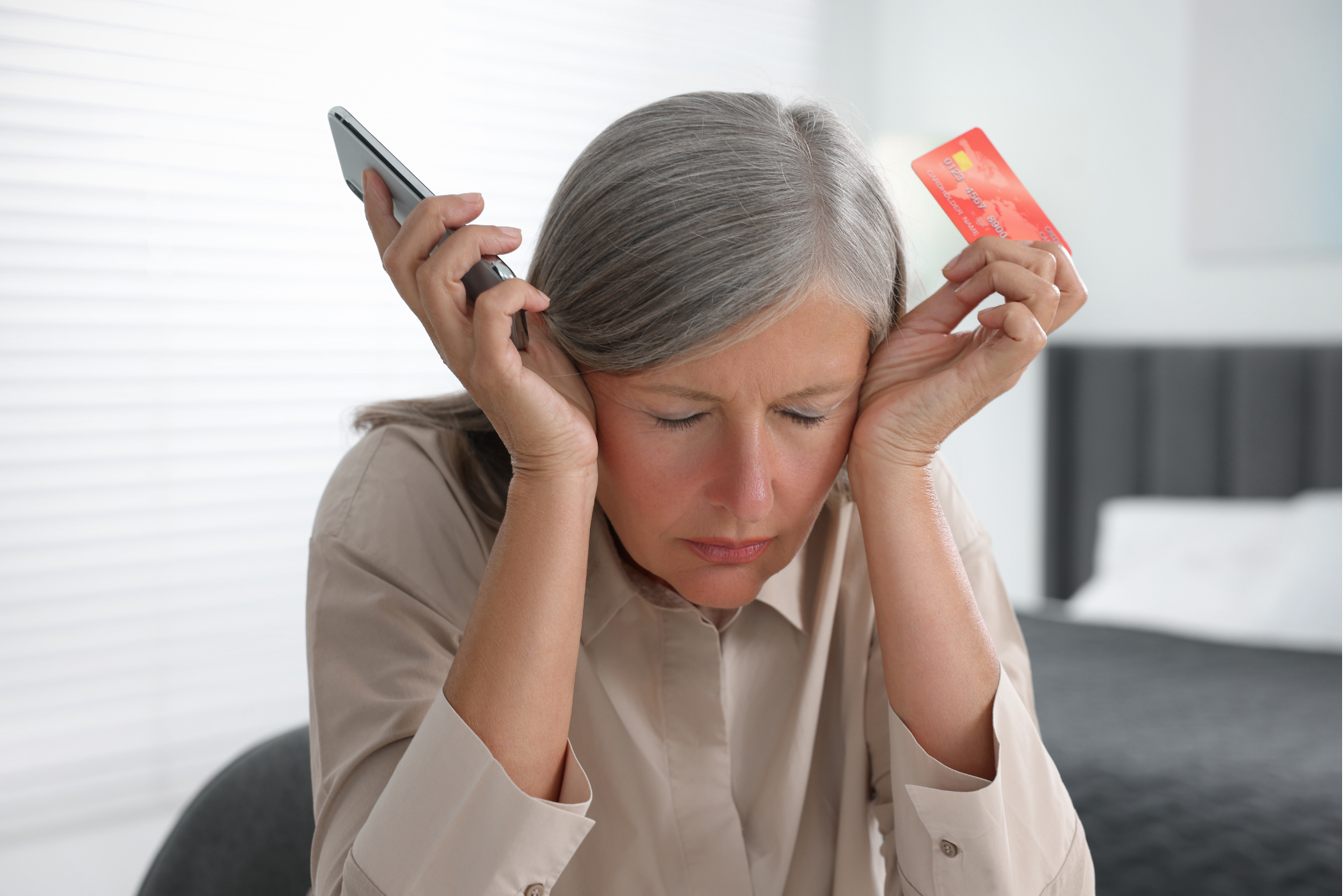 A woman with gray hair looks frustrated while holding a phone and a credit card.