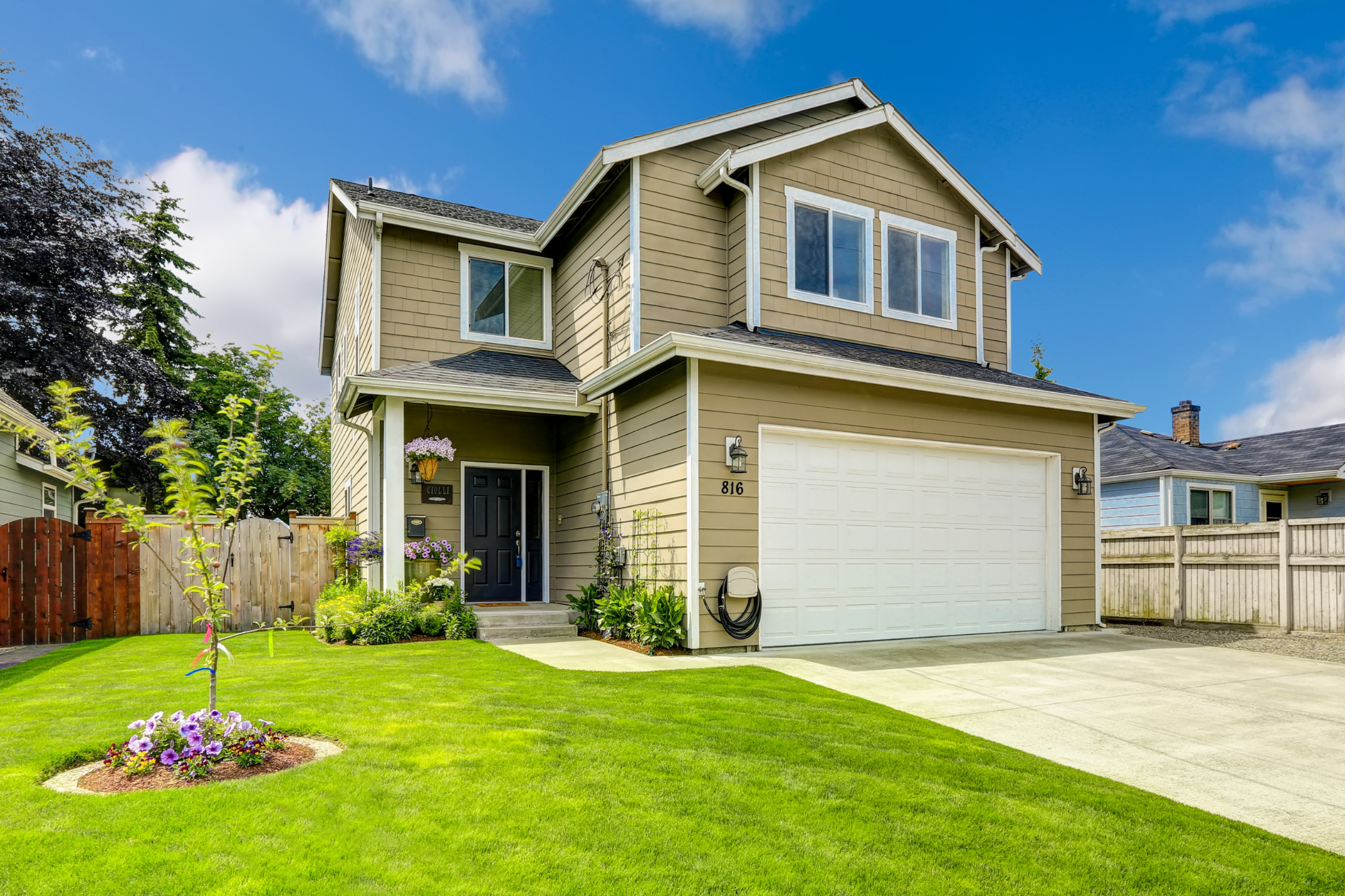 A two-story house with beige siding, white trim, and a large garage. The front yard has a circular flower bed and a well-manicured lawn. The sky is clear and blue.