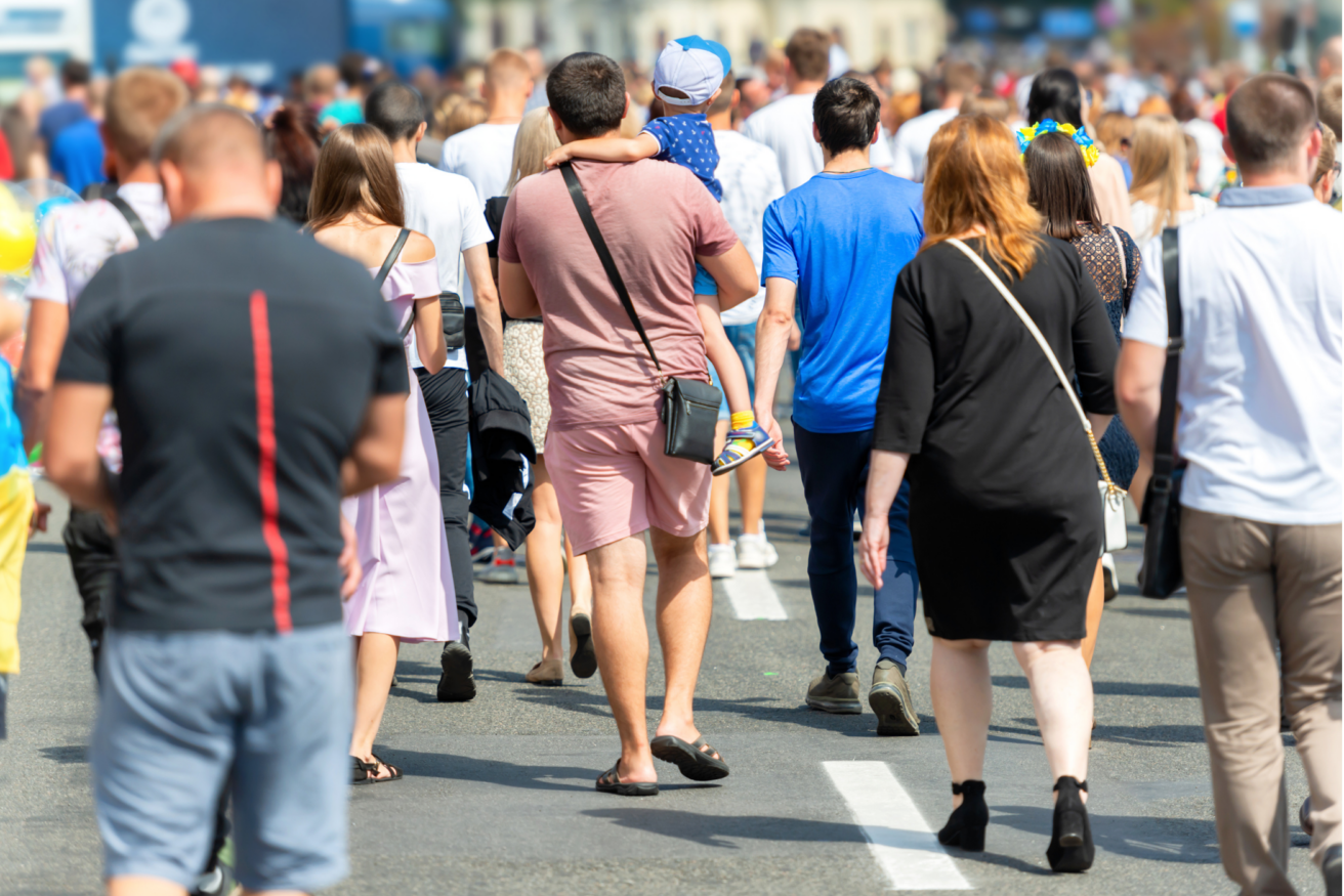 A crowd of people, including a man carrying a child, walk on a sunny street. The group consists of individuals wearing various casual summer clothes.