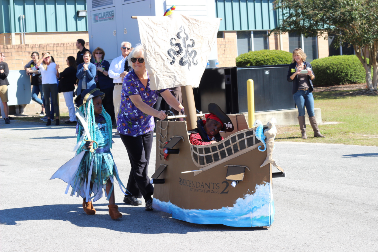 A woman pushes a child in a pirate-themed wheelchair while another child dressed in a costume walks alongside. Several people watch in the background.