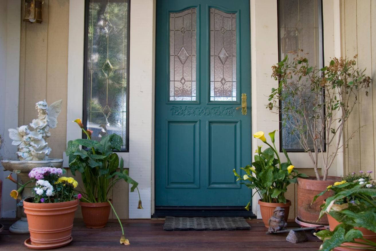 A teal door with decorative glass panels is flanked by two tall windows. Potted plants and flowers are arranged on the wooden porch, along with a frog statue and a white angel sculpture.