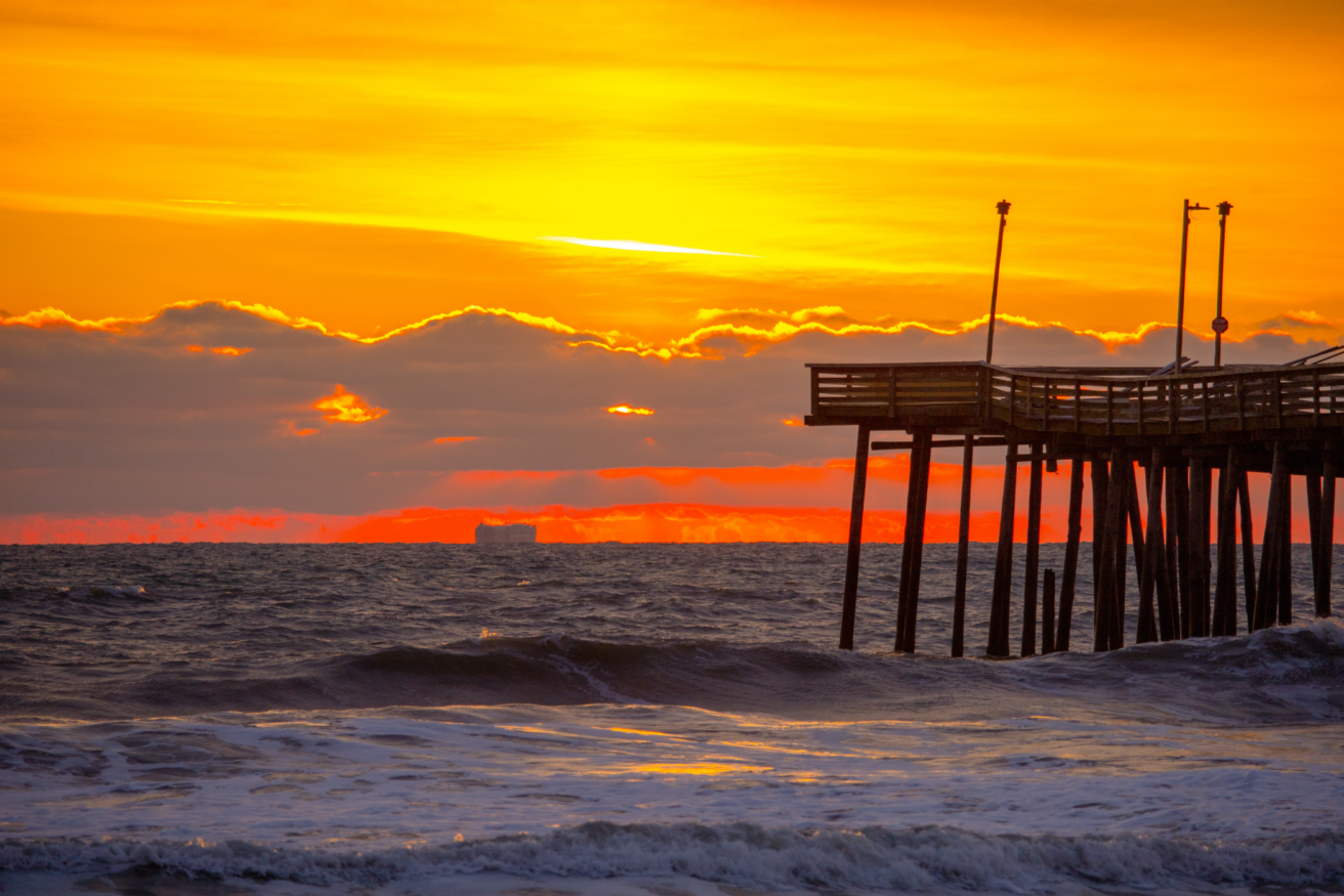 A wooden pier extends over the ocean, backlit by a vibrant orange sunset with waves crashing in the foreground and clouds along the horizon.