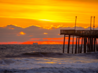 A wooden pier extends over the ocean, backlit by a vibrant orange sunset with waves crashing in the foreground and clouds along the horizon.