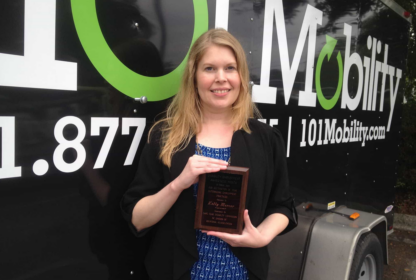 A woman stands in front of a 101 Mobility trailer, holding a plaque.