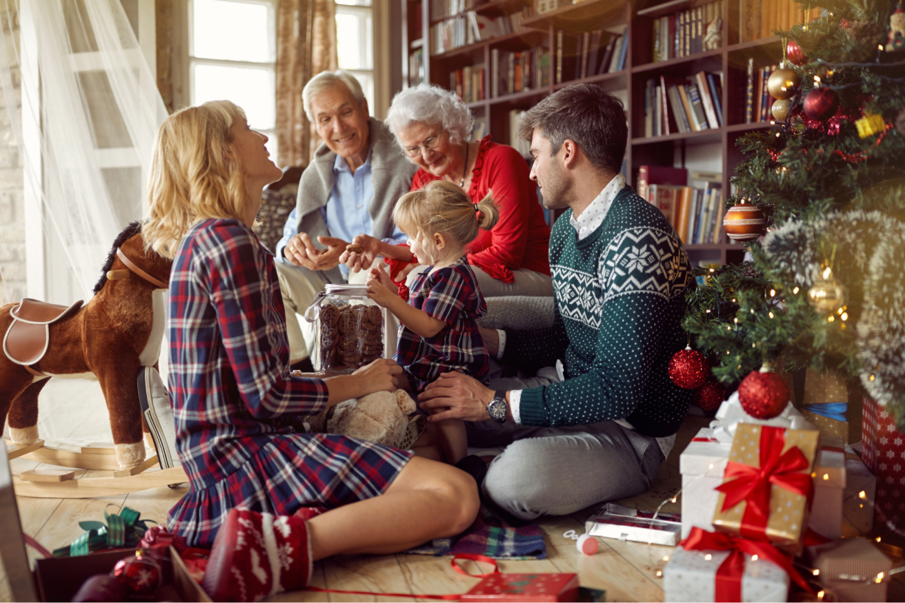 A family is sitting on the floor around a Christmas tree, unwrapping gifts and smiling. The scene includes a decorated tree, wrapped presents, and bookshelves in the background.