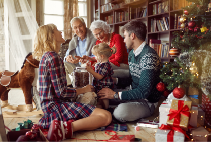 A family is sitting on the floor around a Christmas tree, unwrapping gifts and smiling. The scene includes a decorated tree, wrapped presents, and bookshelves in the background.