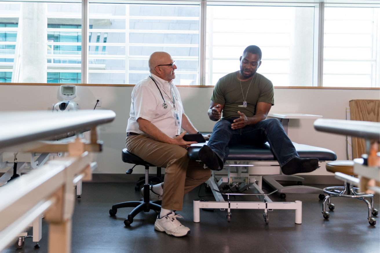 A man in a white coat and stethoscope speaks with a seated man on a medical examination table in a bright, modern clinic.