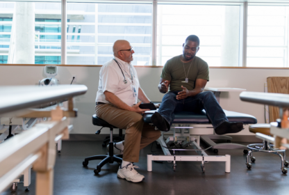 A man in a white coat and stethoscope speaks with a seated man on a medical examination table in a bright, modern clinic.