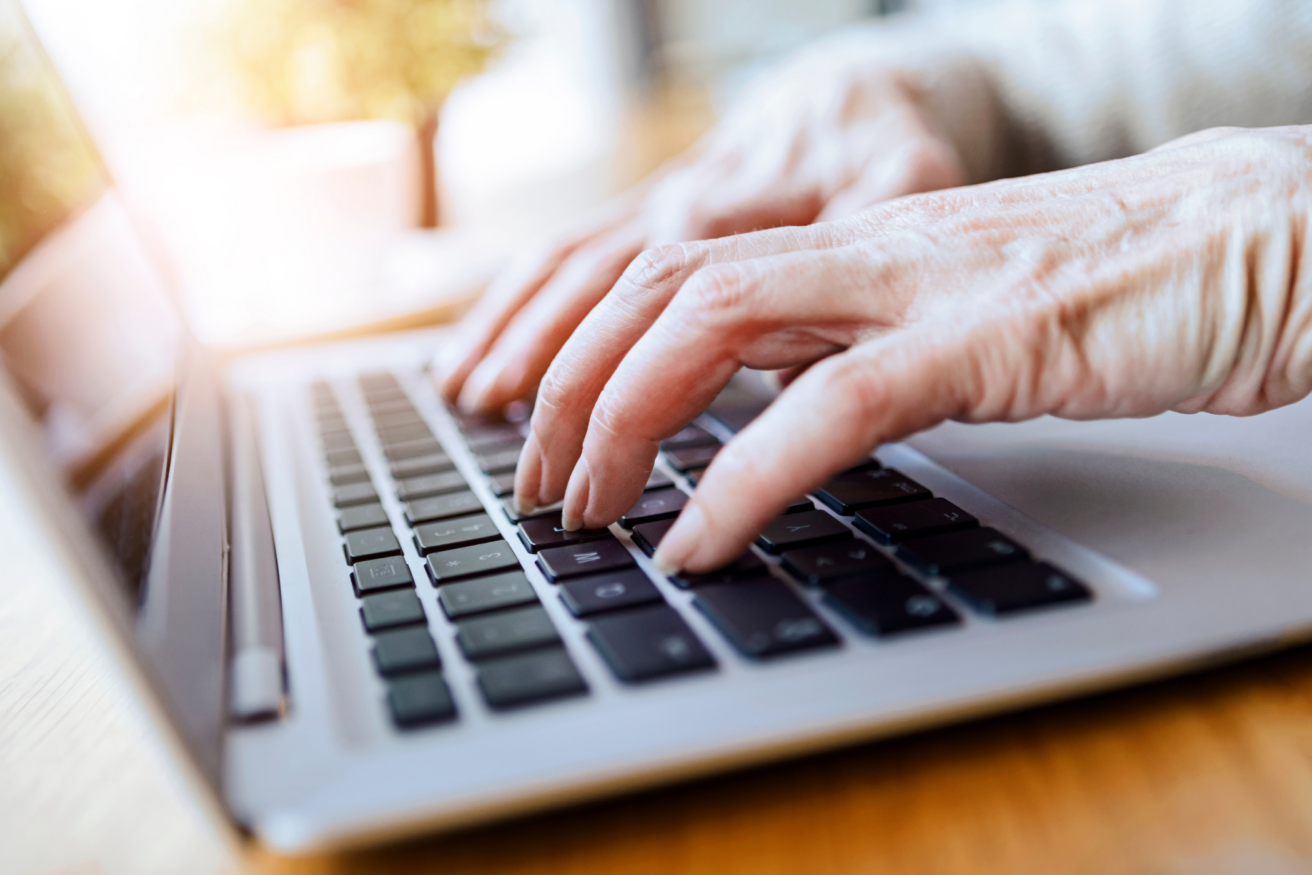 Close-up of an elderly person's hands typing on a laptop keyboard, with sunlight in the background.