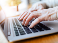 Close-up of an elderly person's hands typing on a laptop keyboard, with sunlight in the background.