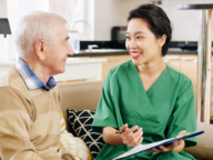 A woman in green scrubs sits on a couch and talks to an older man. She holds a clipboard and a pen.