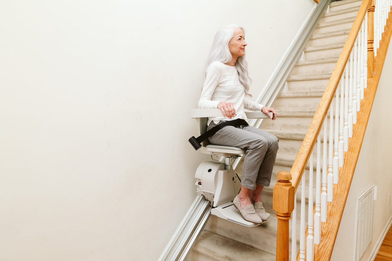 A woman with long white hair rides a stairlift chair up a carpeted staircase next to a wooden railing.