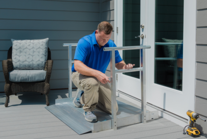 A man in a blue shirt installs a metal wheelchair ramp at the entryway of a home. A wicker chair and a cordless drill are nearby.