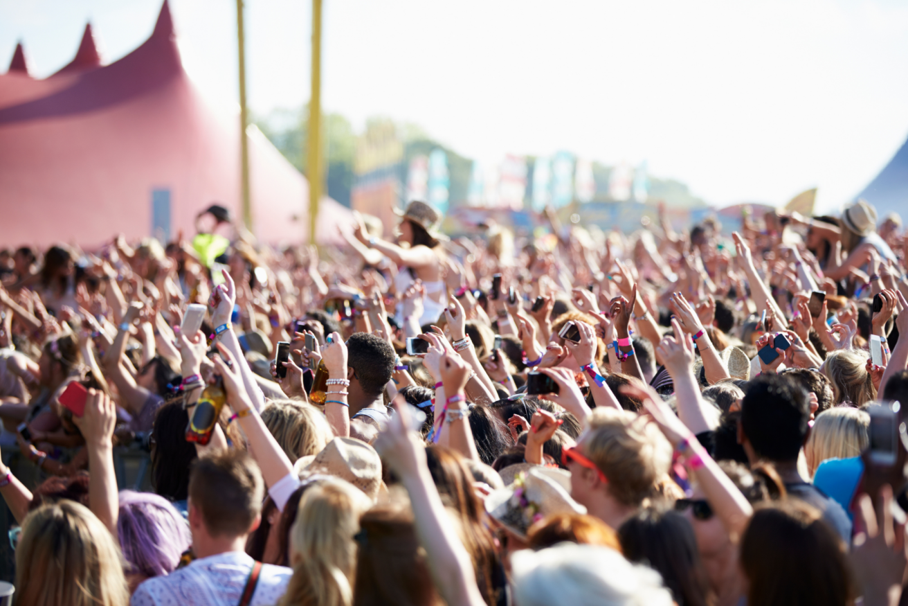 A large crowd of people at an outdoor music festival during the day, many raising their hands and holding smartphones, with large tents visible in the background.