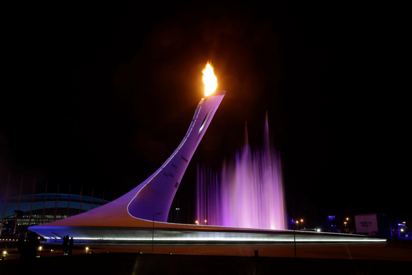 An illuminated torch with a purple-lit water feature at night, possibly part of a major event or sports venue.