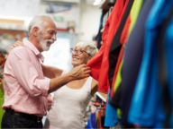 An elderly couple smiles and examines clothing in a store, with colorful garments around them. The man is wearing a pink shirt and the woman a white tank top, both appearing to enjoy their shopping.