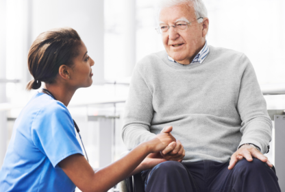 A nurse in blue scrubs holds hands and converses with an elderly man seated in a wheelchair.