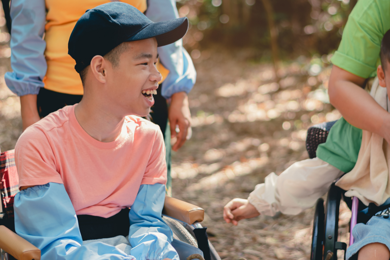 A person in a wheelchair wearing a pink shirt and blue cap smiles outdoors, surrounded by others.