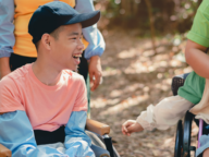 A person in a wheelchair wearing a pink shirt and blue cap smiles outdoors, surrounded by others.