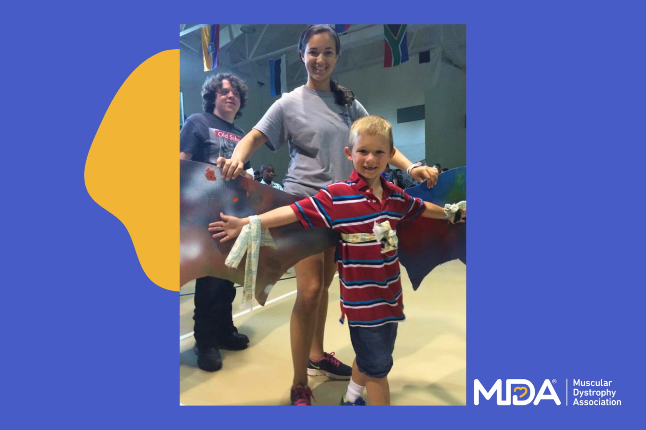 A woman and a young boy, both smiling, hold a large bat-shaped cutout. Another person stands in the background. MDA logo and "Muscular Dystrophy Association" text in the bottom right corner.