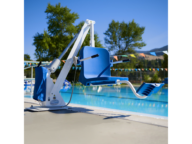 A blue pool lift chair positioned at the edge of an outdoor swimming pool on a sunny day. Trees and colorful flags are visible in the background.