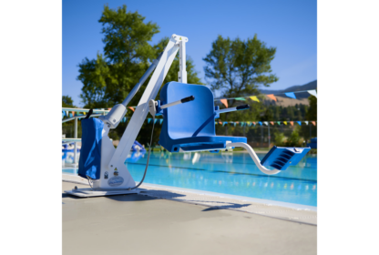 A blue pool lift chair positioned at the edge of an outdoor swimming pool on a sunny day. Trees and colorful flags are visible in the background.