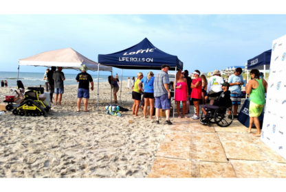 A group of people stands under tents on a sandy beach, some wearing T-shirts and hats. A person in a wheelchair is also present, with a beach and ocean in the background.