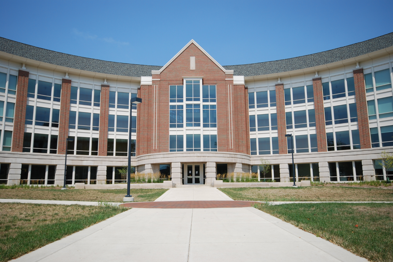 A modern brick and glass building with a curved facade and a central entrance, surrounded by grass and a paved walkway under a clear blue sky.