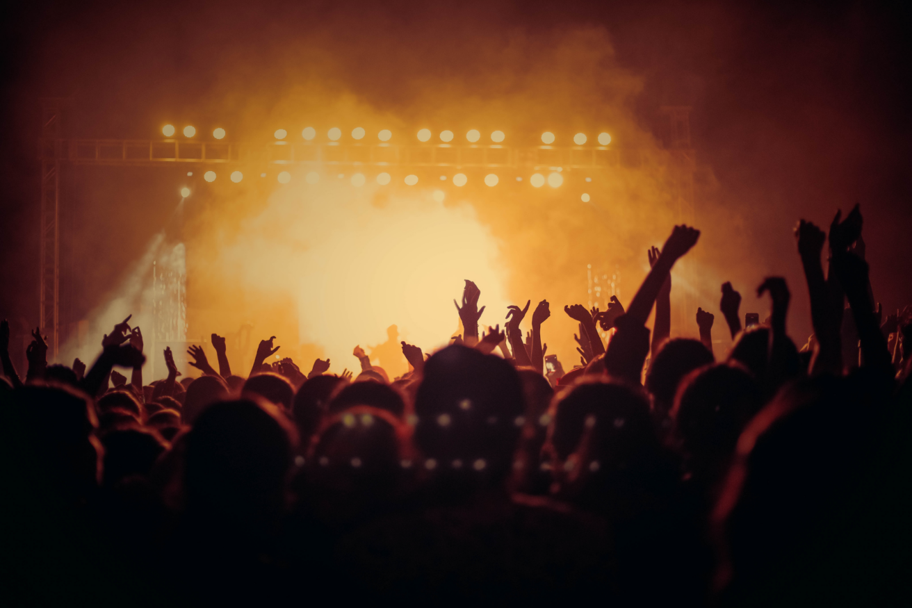 A crowd of people with raised hands at a concert, illuminated by bright stage lights and a hazy atmosphere.