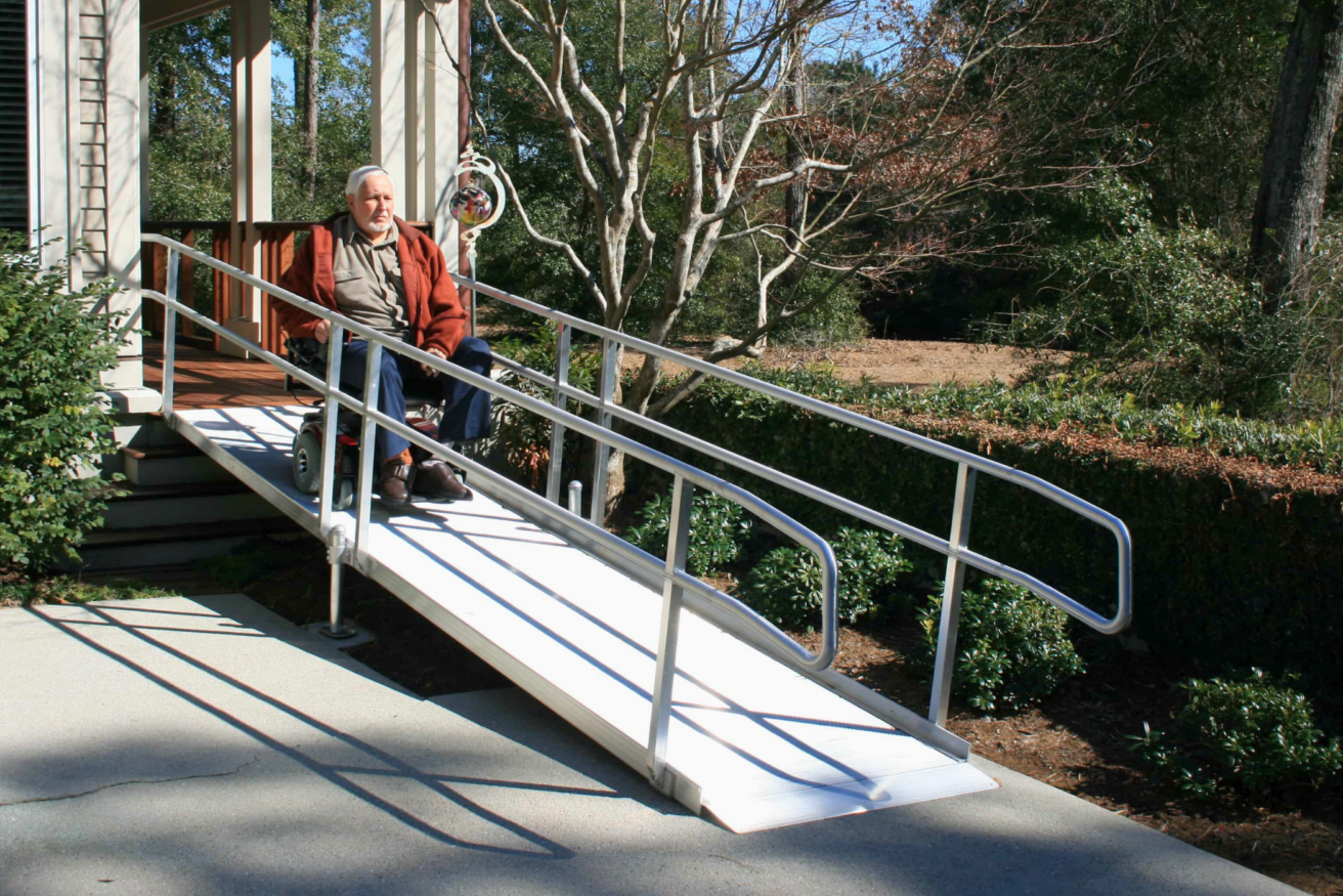 A person in a motorized wheelchair uses a ramp to enter a house with a covered porch and large windows, surrounded by trees and shrubs.