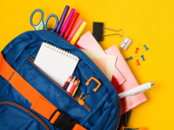 A blue backpack with orange accents contains colorful pens, a notepad, scissors, papers, a pen, a binder clip, a sharpener, and push pins, all set against a yellow background.