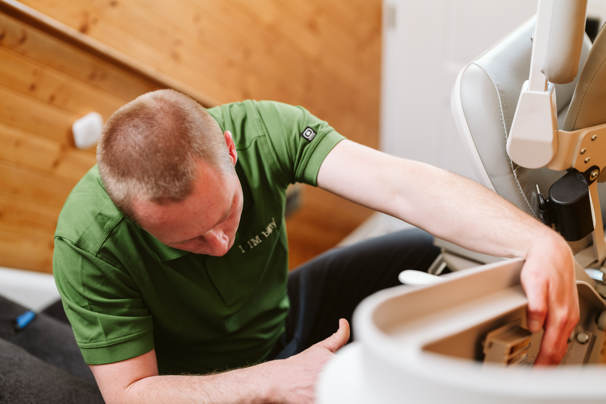 A person in a green shirt is kneeling and working on a stairlift chair, adjusting a component.