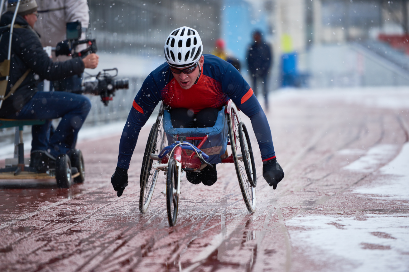 A person in a racing wheelchair competes on a snowy track, wearing a helmet and winter athletic gear. A cameraman is filming to the side.