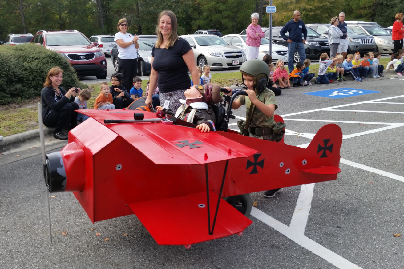 A child in military gear pushes a wheelchair-bound person in a red airplane costume; a woman stands nearby. Crowds in the background watch and take photos in a parking lot.