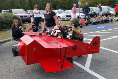 A child in military gear pushes a wheelchair-bound person in a red airplane costume; a woman stands nearby. Crowds in the background watch and take photos in a parking lot.