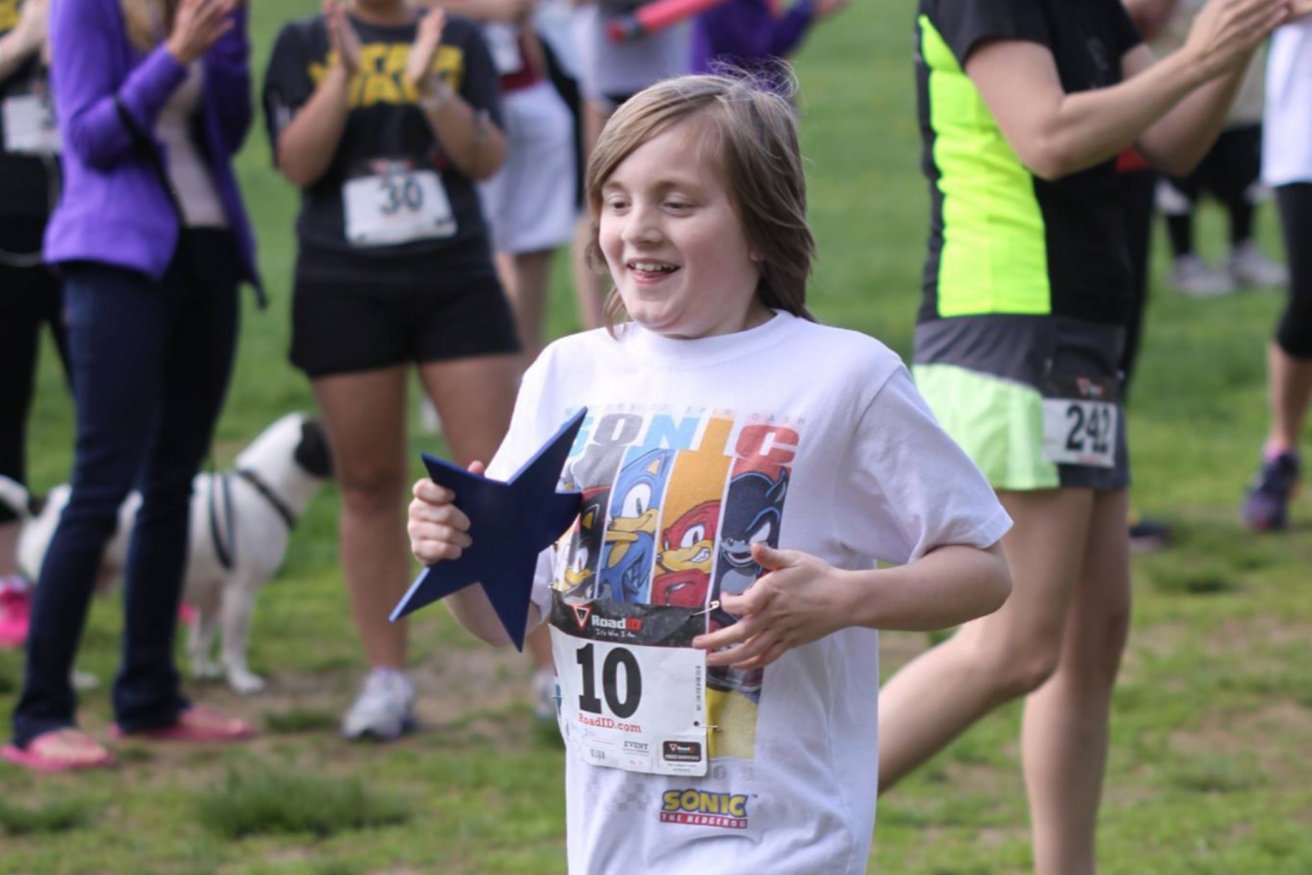 A young child holding a blue star-shaped object runs in a park, wearing a Sonic the Hedgehog T-shirt and a race number 10 bib. Other participants and spectators are in the background.