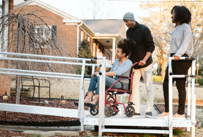 A person in a wheelchair is being pushed up a ramp by another individual. Another person stands beside them, smiling. A brick house is in the background.