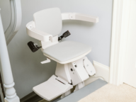 A stairlift is installed on a staircase, featuring a padded seat, backrest, and safety arms.