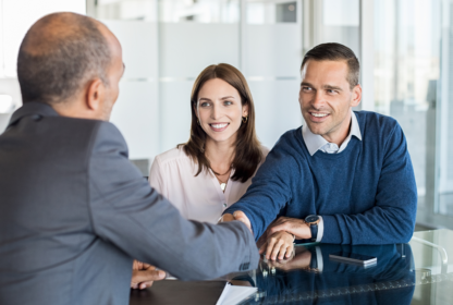 Two people, a woman and a man, smiling and sitting at a table while the man shakes hands with another person in a suit in an office setting.