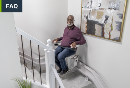 A man sits on a stair lift installed on a curved staircase in a residential setting. He is smiling and holding the control switch, with modern wall decor in the background.