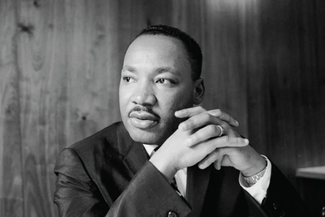 A man in a suit sits with his hands clasped, looking thoughtfully to the side. The background features wood paneling.