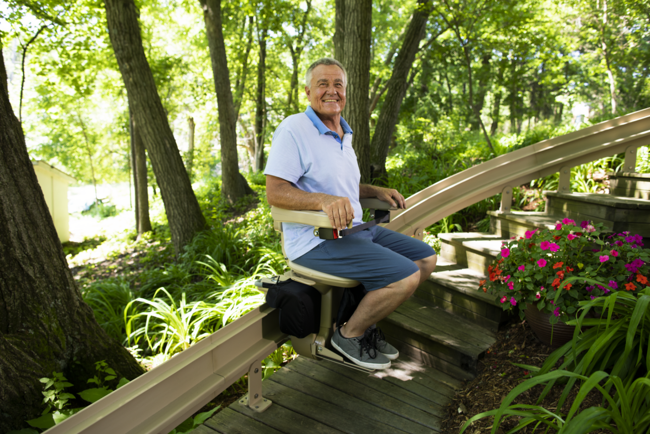 A man in a light blue polo shirt and shorts smiles while sitting on an outdoor stair lift surrounded by greenery and flowers.