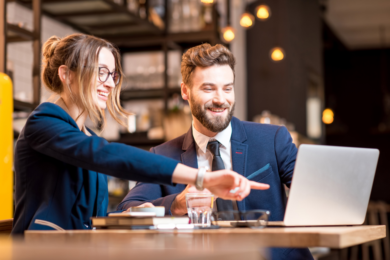 Two business professionals in a cafe setting working together on a laptop, smiling and discussing something on the screen. The woman is pointing at the laptop while the man looks on.